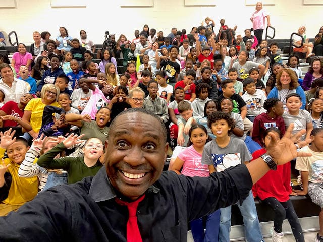 Jamahl Keyes leading a Red Ribbon Week assembly with students raising red wrist ribbons in a school gym in Indianapolis 