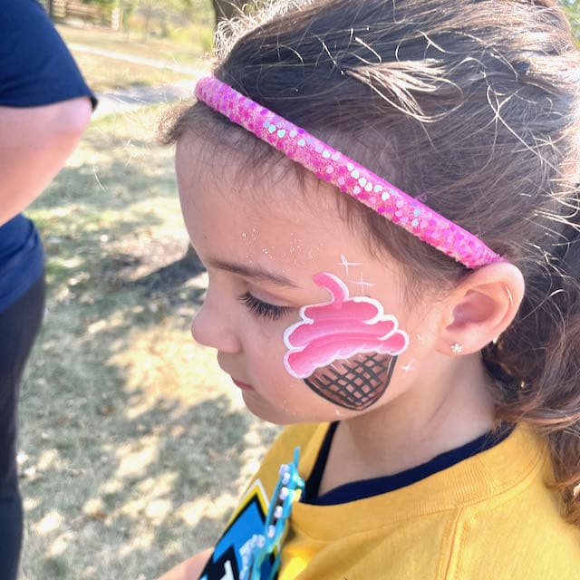 Greenwood face painting artist applying a glittery flower design to a child’s face at a local park event.