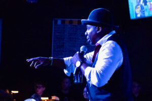 Male singer on stage in a hat and vest, holding a microphone and pointing to the crowd under blue lighting.